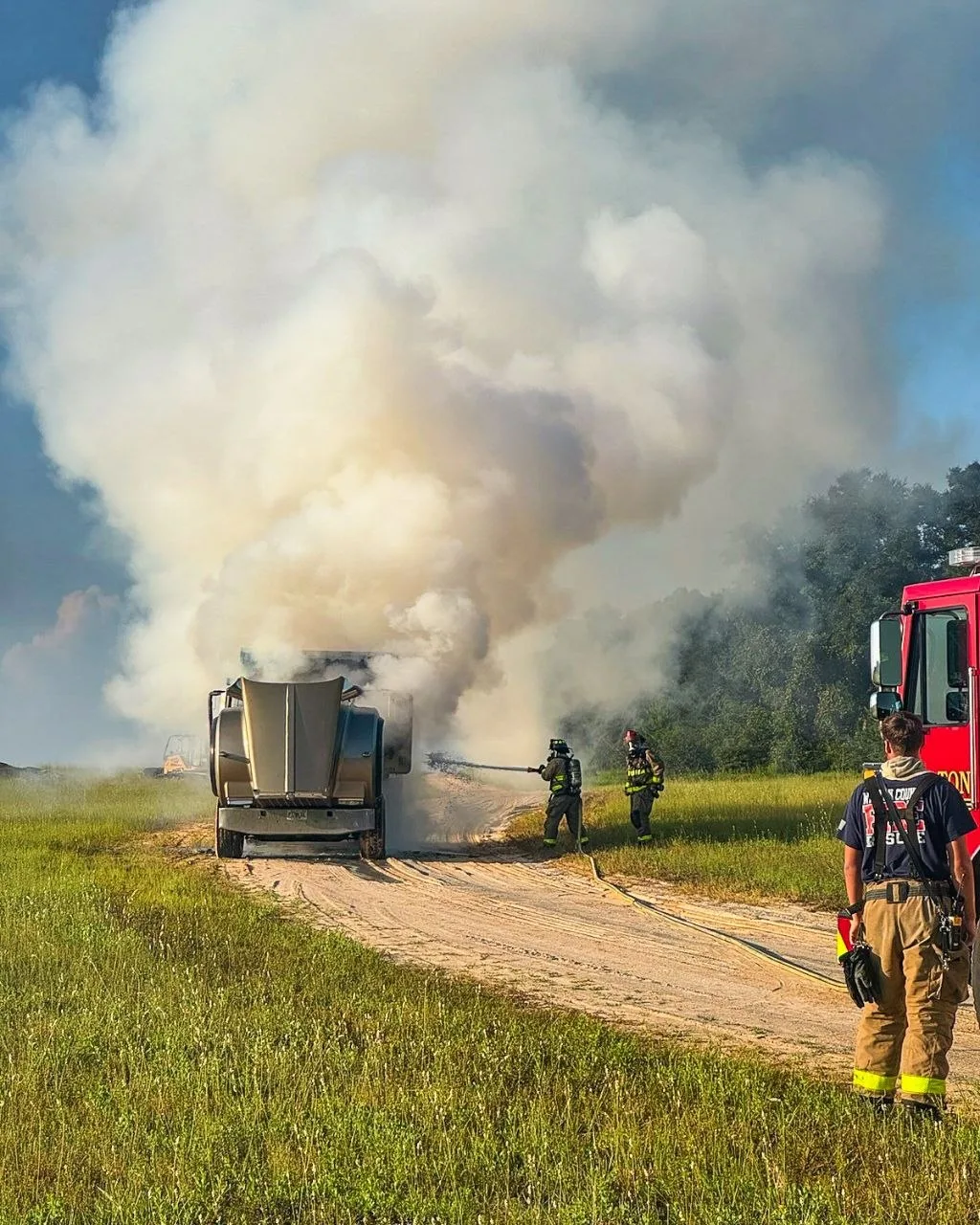 Firefighters direct a hose stream at a burning dump truck as heavy smoke billows over a rural Freeport road