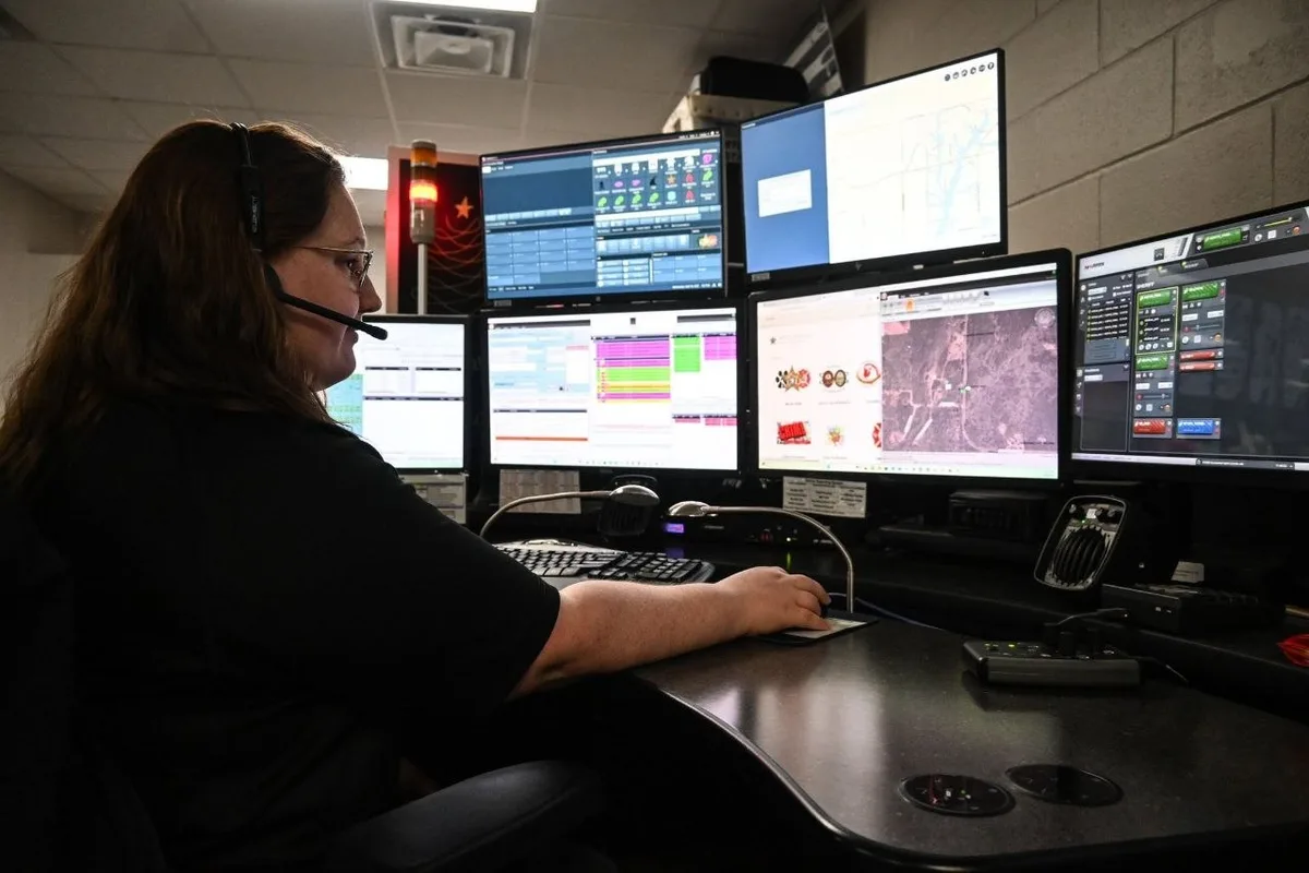 A dispatcher wearing a communications headset monitors a bank of six screens inside a dimly lit emergency communications center